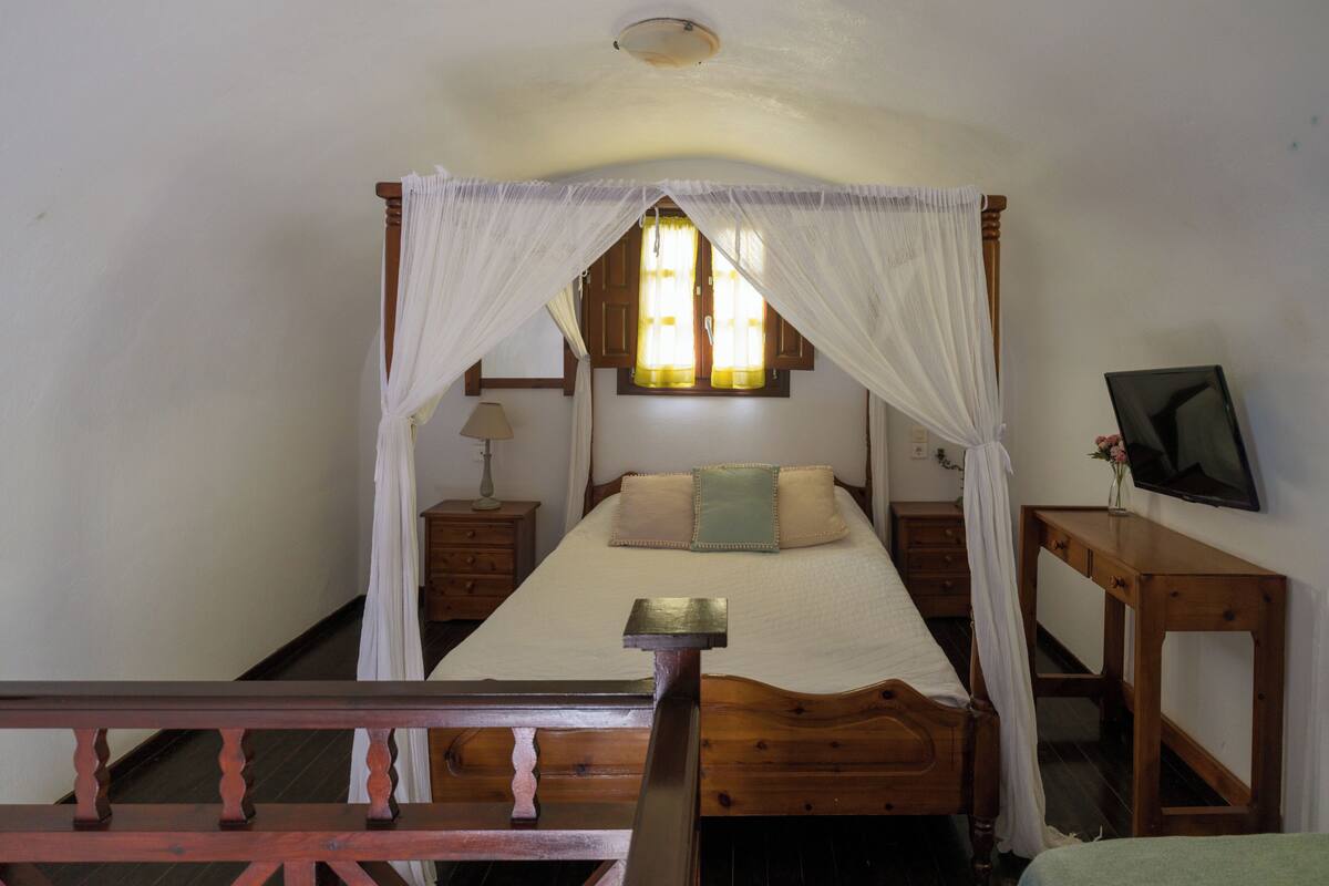 A bedroom with a four-poster bed, a bedside table, a lamp, a TV, and a window with curtains.