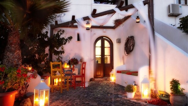 A cobblestone pathway leading to a white building with a wooden door and windows. There are chairs and tables set up outside, and lanterns are placed along the path. - Anna Traditional Apartments (Santorini)