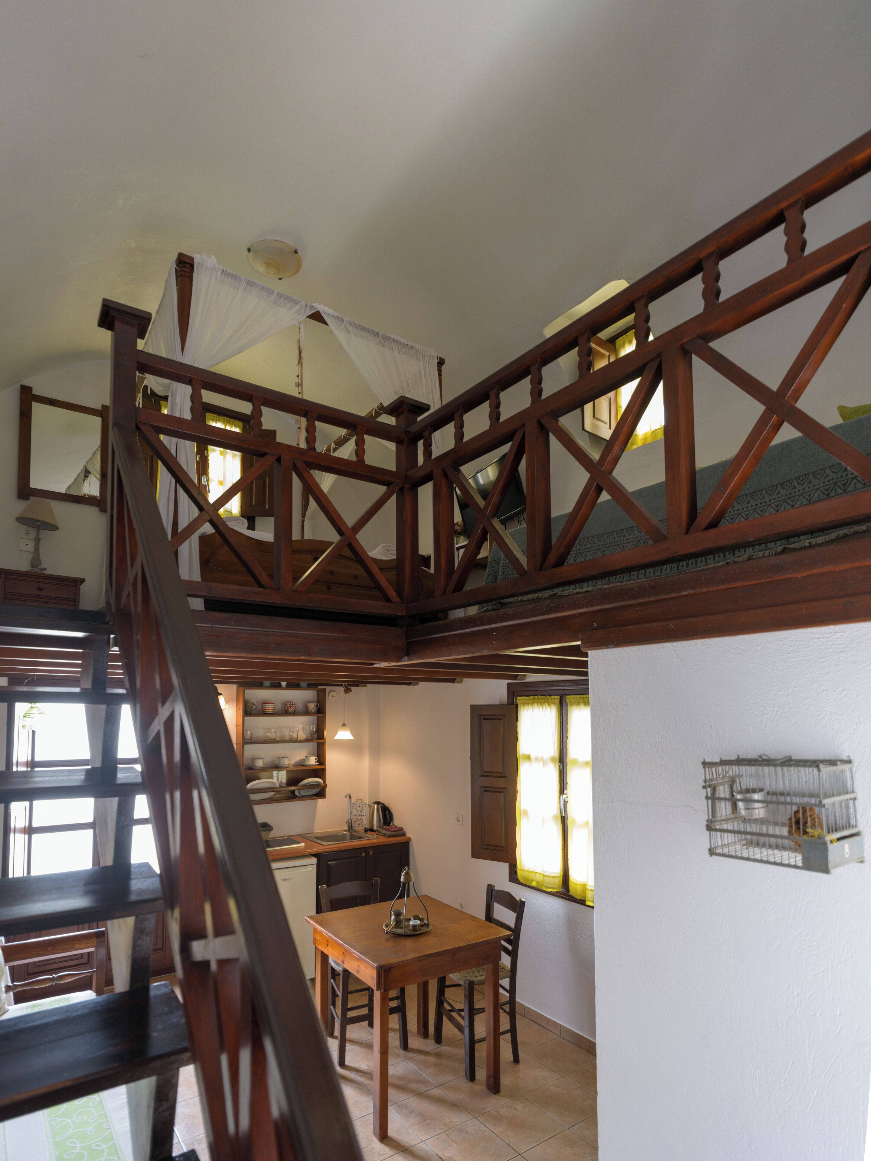 A wooden staircase leading to a loft bedroom with a canopy bed, a dining area with a table and chairs, and a kitchen area with a sink and cabinets.