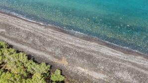 An aerial view of a beach with clear water, pebbly shore, and dense green vegetation. - Anna Traditional Apartments (Santorini)
