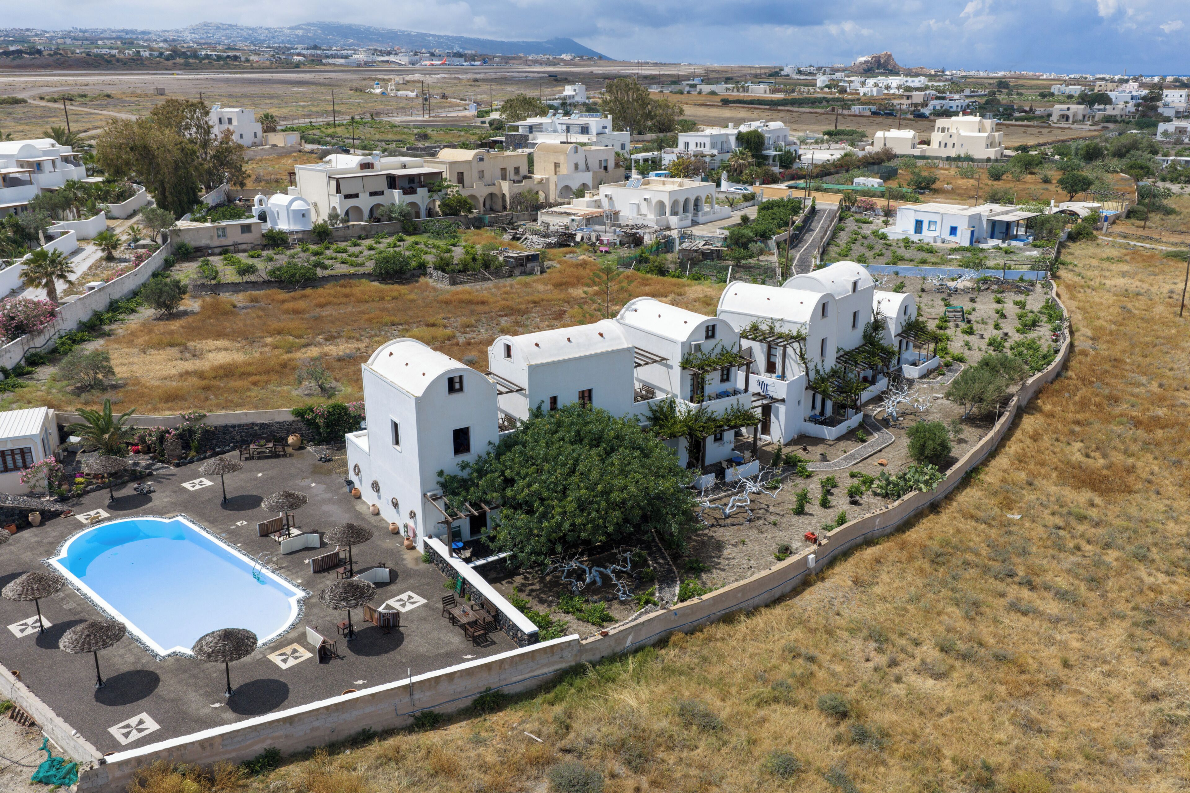 A white building with a swimming pool, surrounded by dry grass and other buildings.