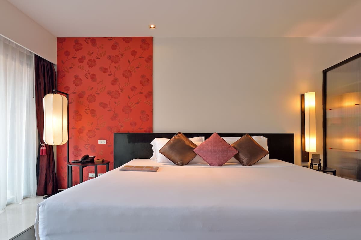 A hotel room with a large bed, a bedside table with a lamp, and a view of the outside through sheer curtains.