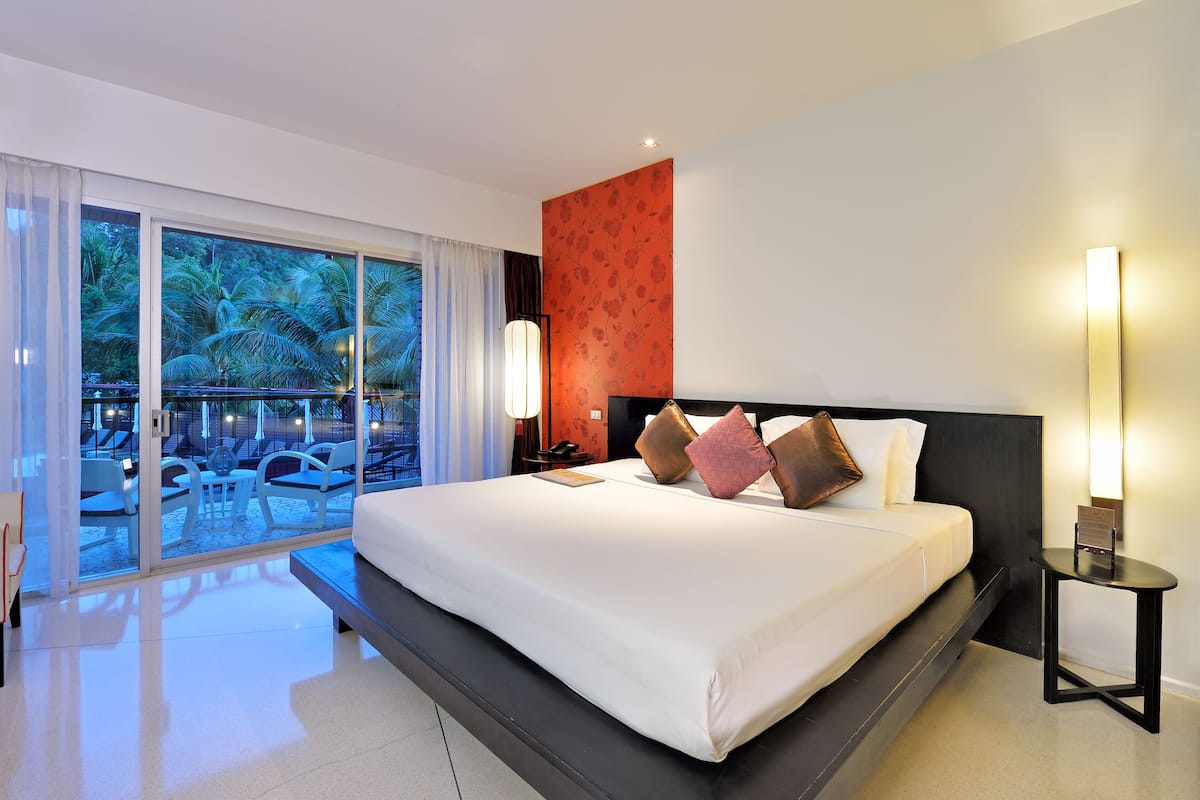 A modern hotel room with a large bed, a nightstand, a lamp, and a view of a pool and palm trees through the glass door.