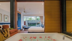 A hotel room with a large bathtub filled with water and rose petals, a bed, a seating area with a table and chairs, and a view of the outdoors through a large window.