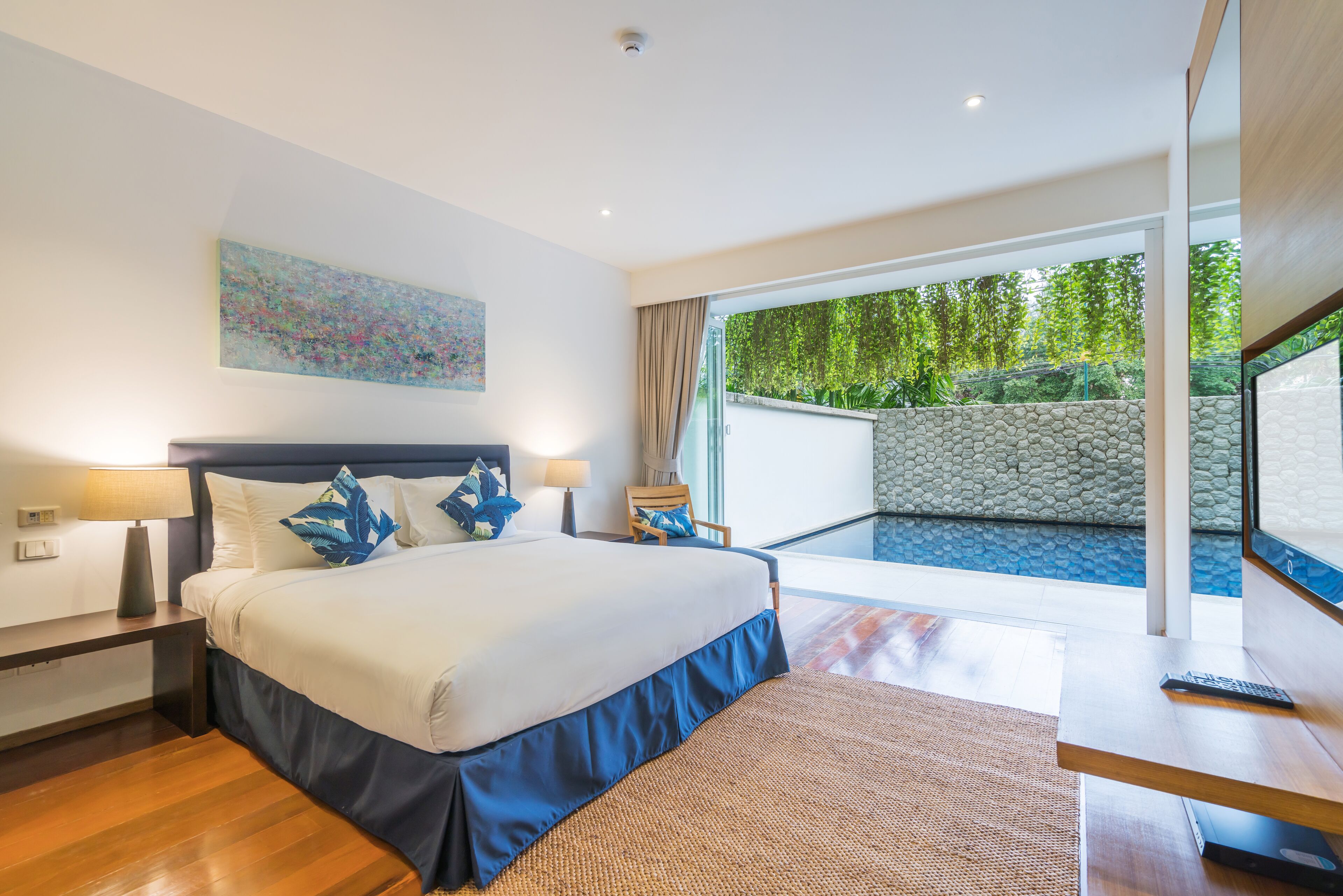 Una habitación de hotel moderna con una cama grande, un escritorio, una computadora portátil y vistas a una piscina y vegetación.