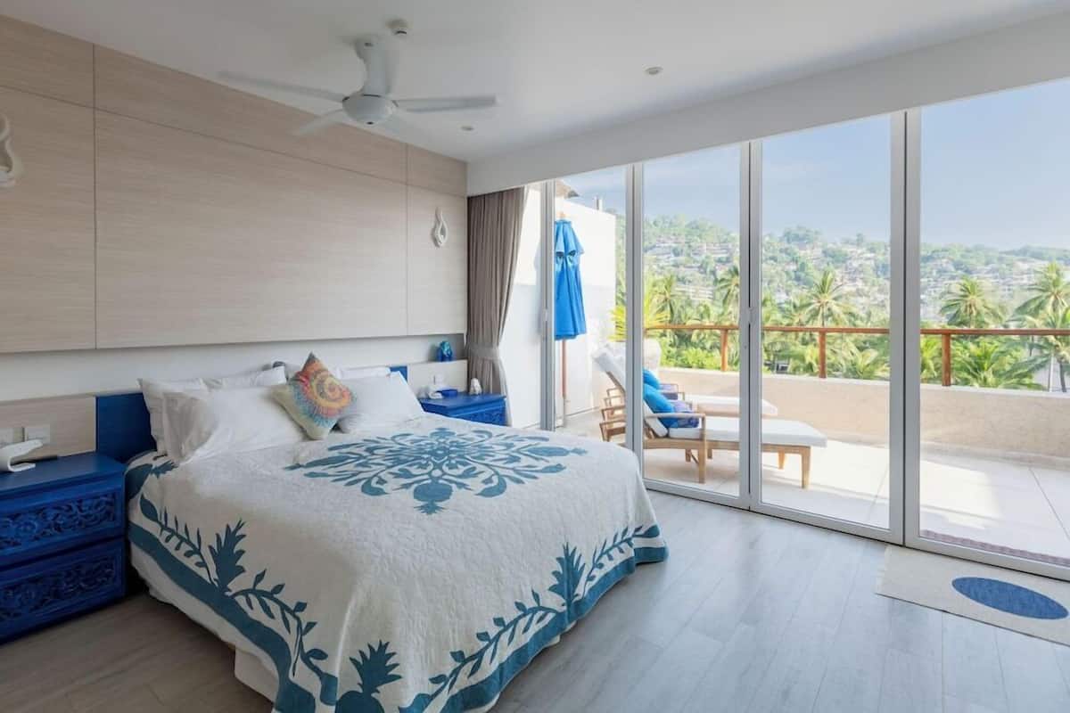 A bedroom with a large bed, a nightstand, a ceiling fan, and a balcony with a view of palm trees and a cityscape.