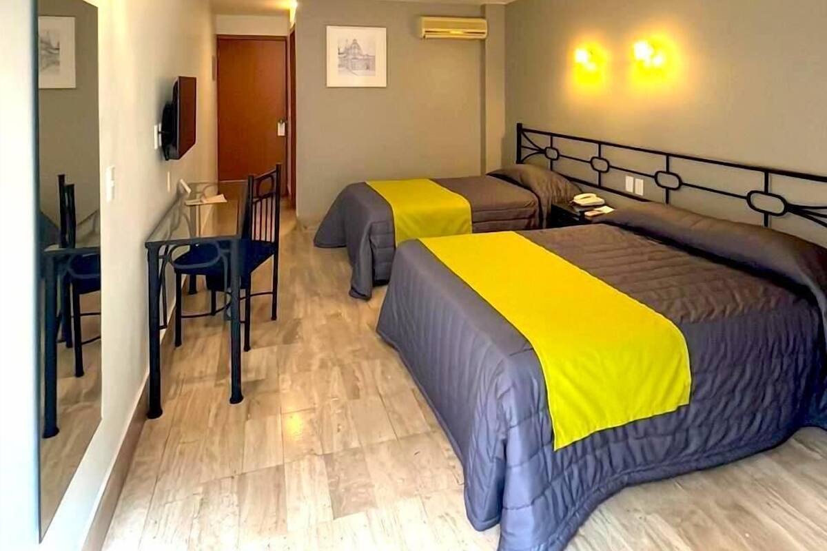 A hotel room with two beds, a desk with a chair, a mirror, and an air conditioning unit.