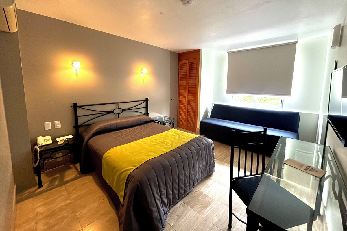 A hotel room with a bed, bedside table, lamp, and a seating area with a view of the outdoors.