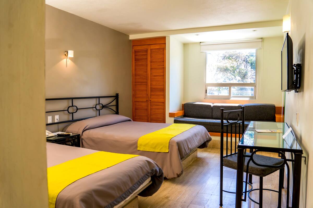 A hotel room with two beds, a desk, a chair, a sofa, and a window with a view.