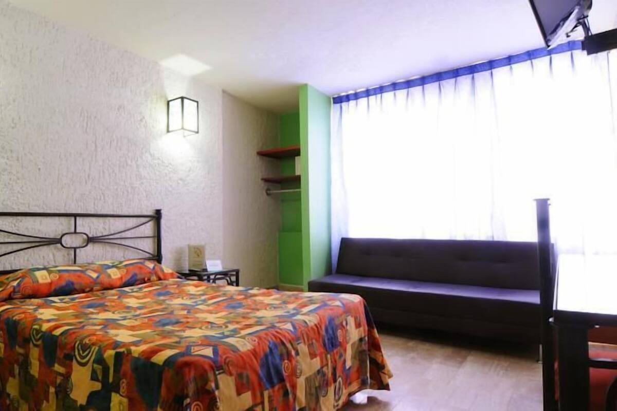 A bedroom with a bed, a bedside table, a lamp, a sofa, and a window with curtains.