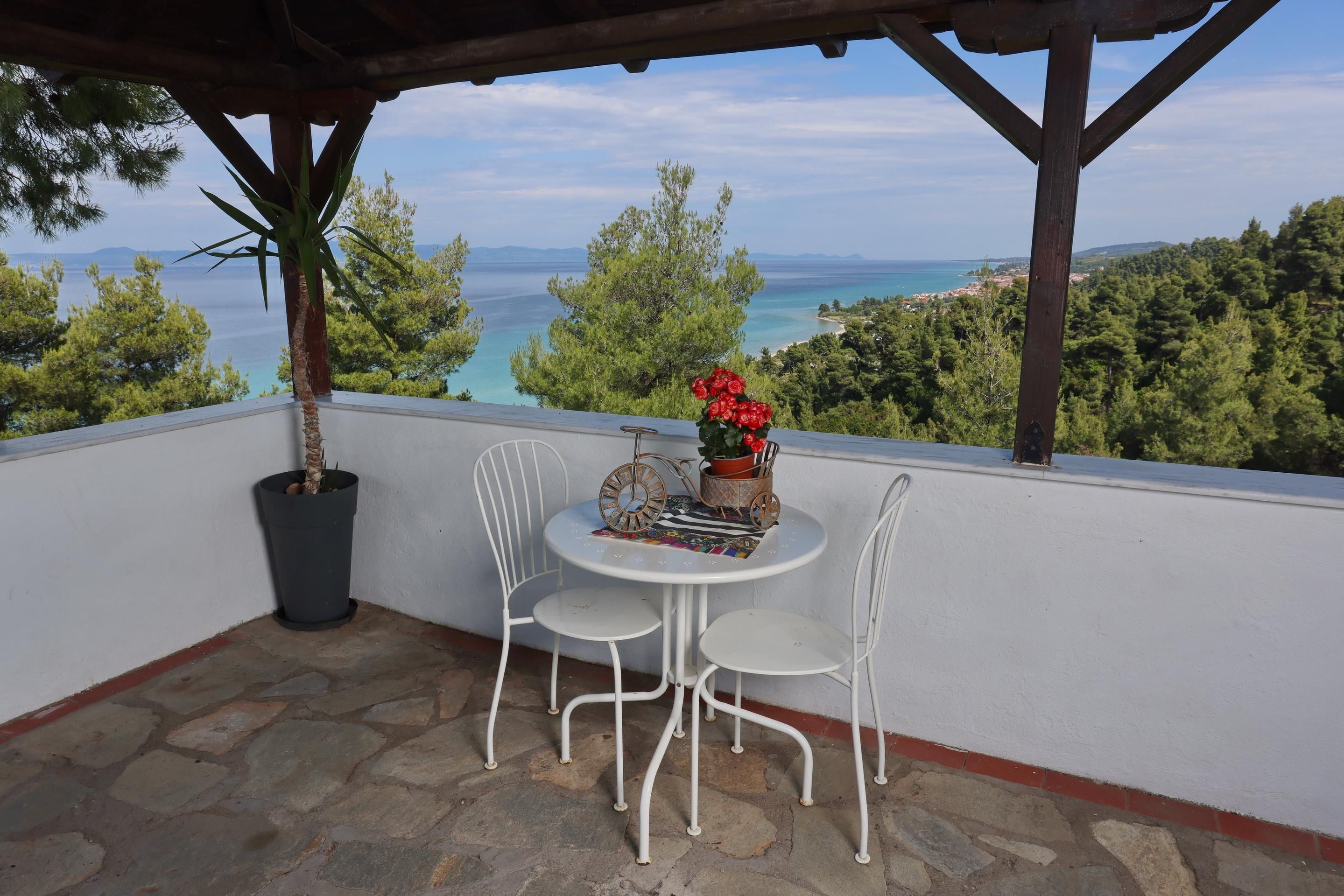 A balcony with a table and chairs, overlooking a coastal landscape with trees and a view of the sea.