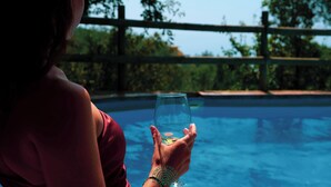 Una persona sosteniendo un vaso de agua junto a una piscina, con una valla de madera y árboles al fondo.