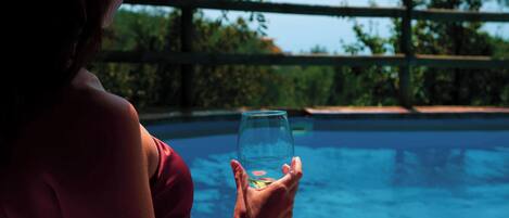 A person holding a glass of water by a pool with a wooden fence and trees in the background.