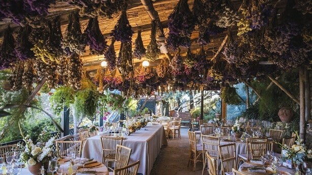 Un área de comedor al aire libre con mesas dispuestas para una comida, rodeada de arreglos florales colgantes.