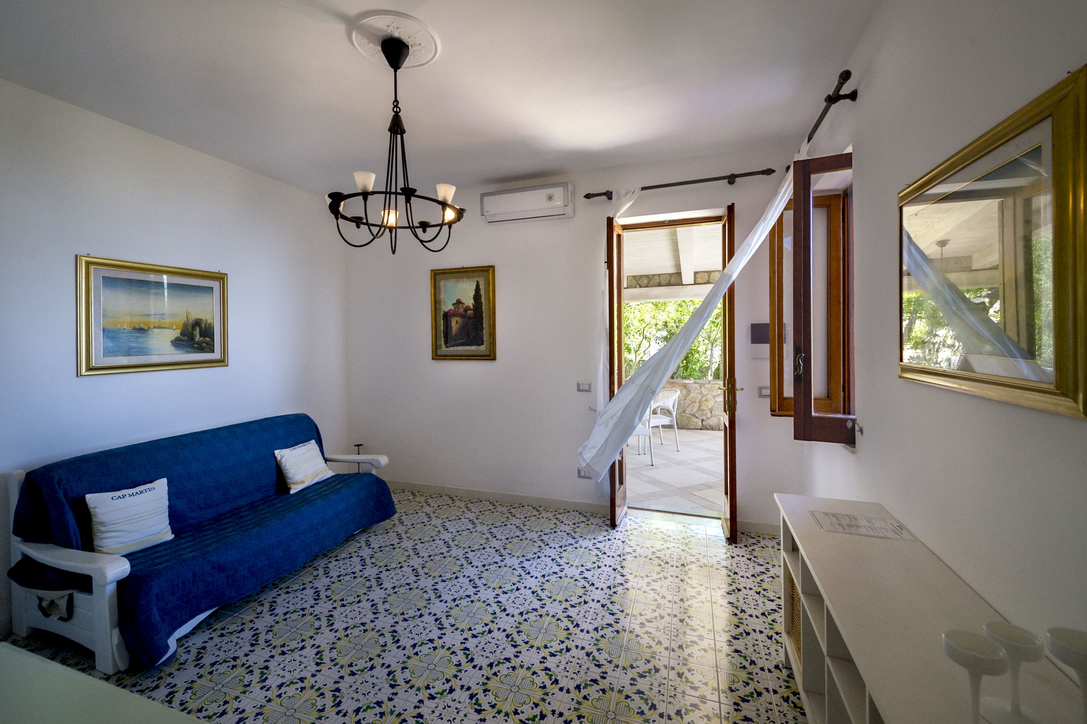 Una habitación con un sofá azul, una araña de techo, dos cuadros enmarcados, una escalera y un ventanal con vistas.