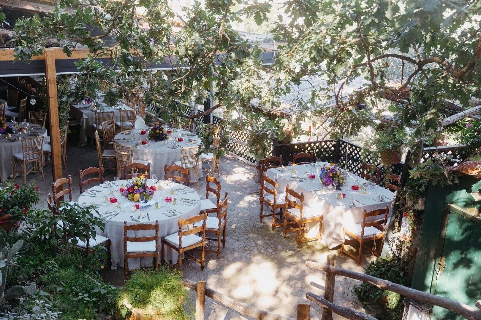 An outdoor dining area with round tables and wooden chairs, surrounded by greenery and a wooden pergola.