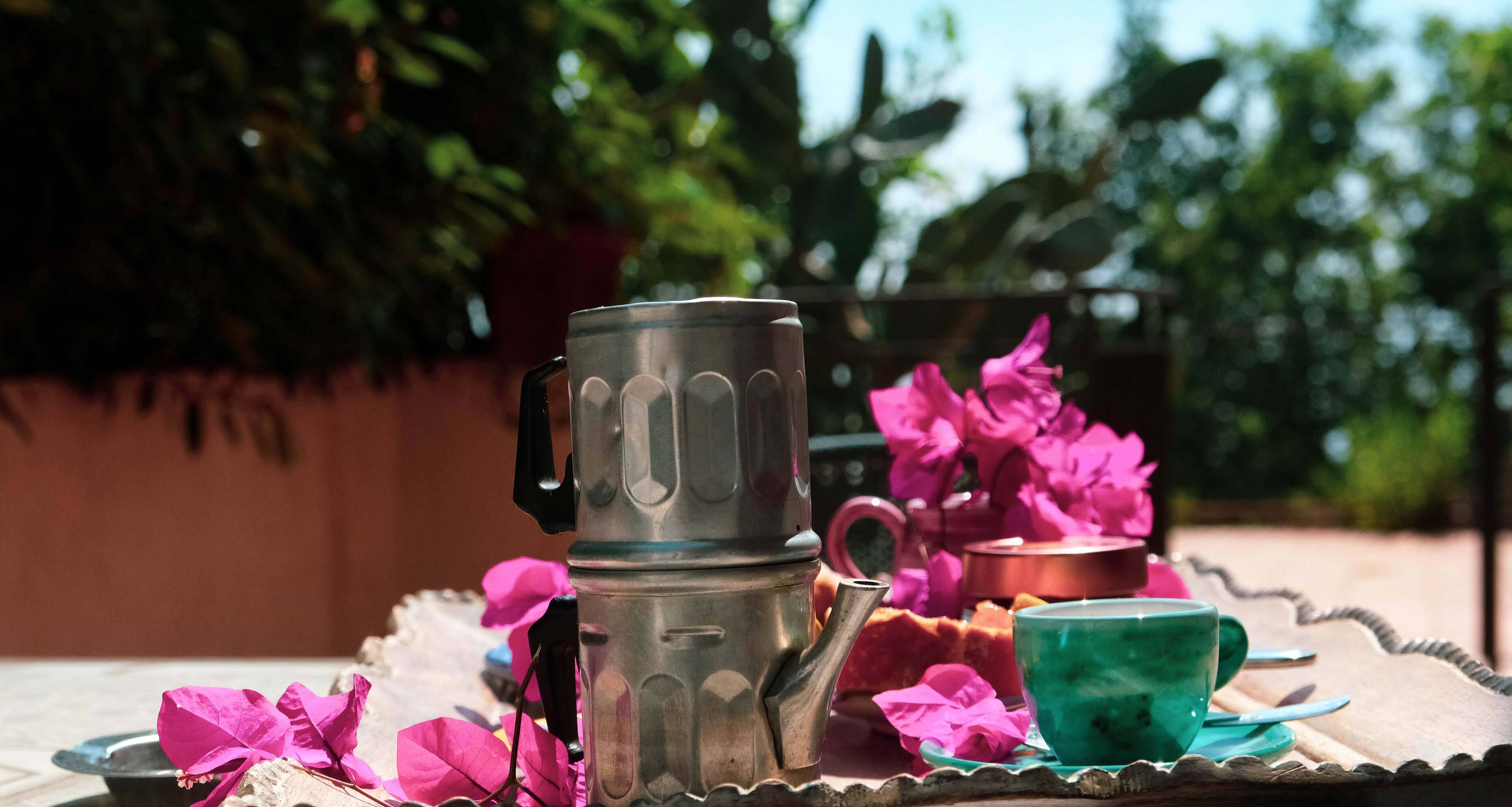 Eine Metall-Kaffeekanne, eine grüne Tasse und rosa Blumen auf einem Tisch im Freien.