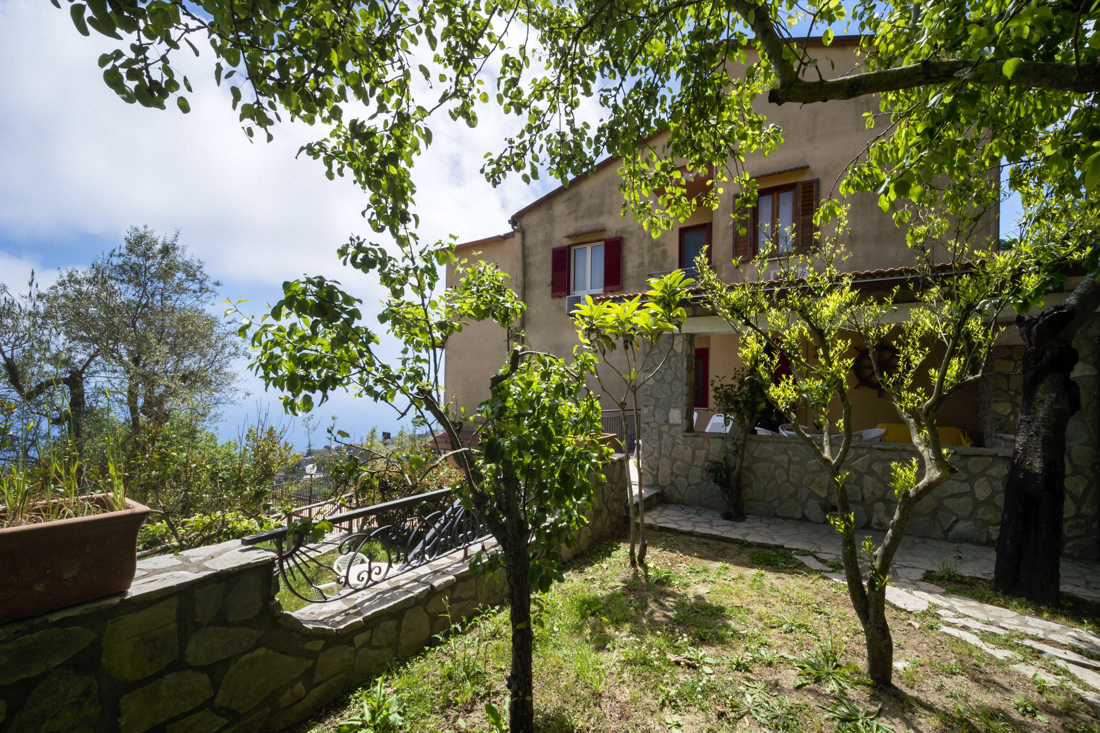 Une cour entourée de murs en pierre, avec un petit arbre, une rambarde en métal et un bâtiment aux volets rouges et doté d’un balcon.