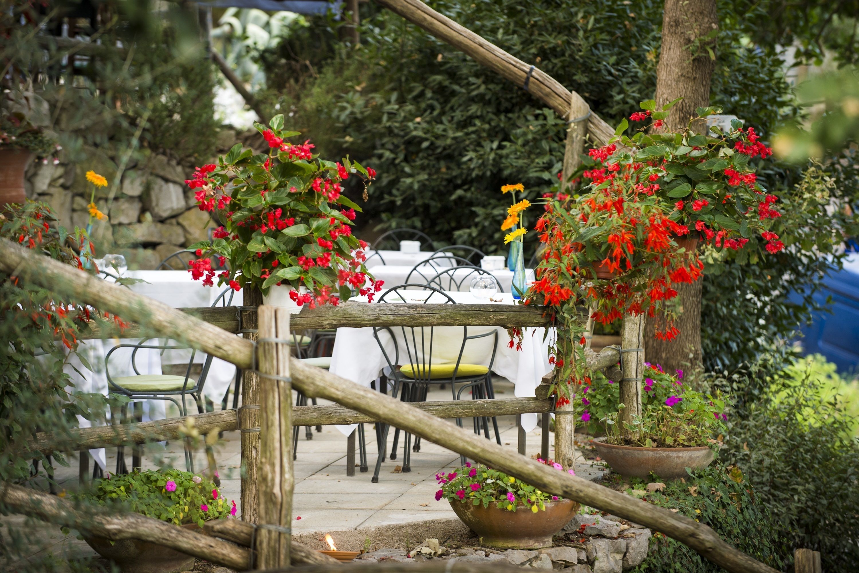 A rustic outdoor seating area with wooden tables and chairs, surrounded by potted plants and hanging flower baskets.