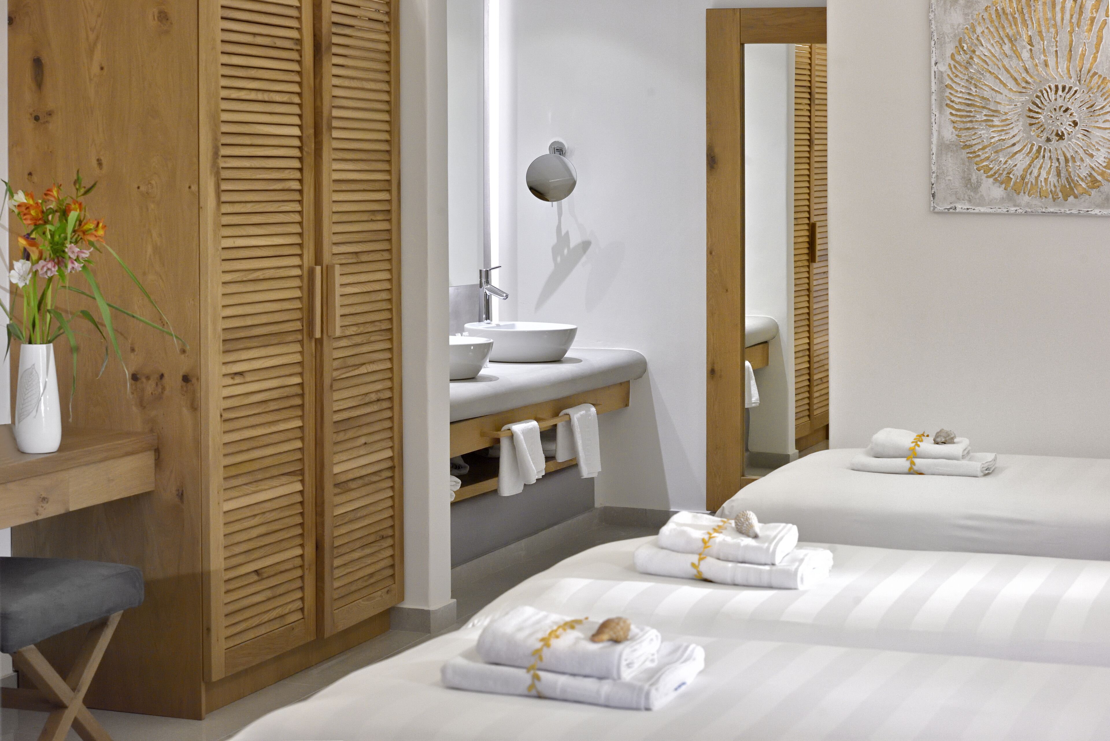 A modern bedroom with a wooden wardrobe, a bedside table with a vase of flowers, a sink, and two beds with towels.