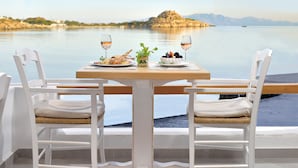 A dining table with two white chairs, a plate of seafood, a glass of wine, and a view of a calm lake with mountains in the background. - Hotel Petinos (Mykonos)