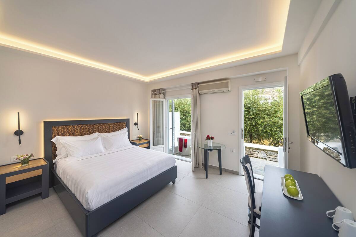 A modern hotel room with a large bed, bedside tables, a small table with a vase, and a TV on the wall.