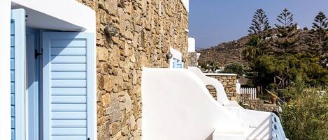 A balcony with a stone wall, a white building, and a view of a hillside.