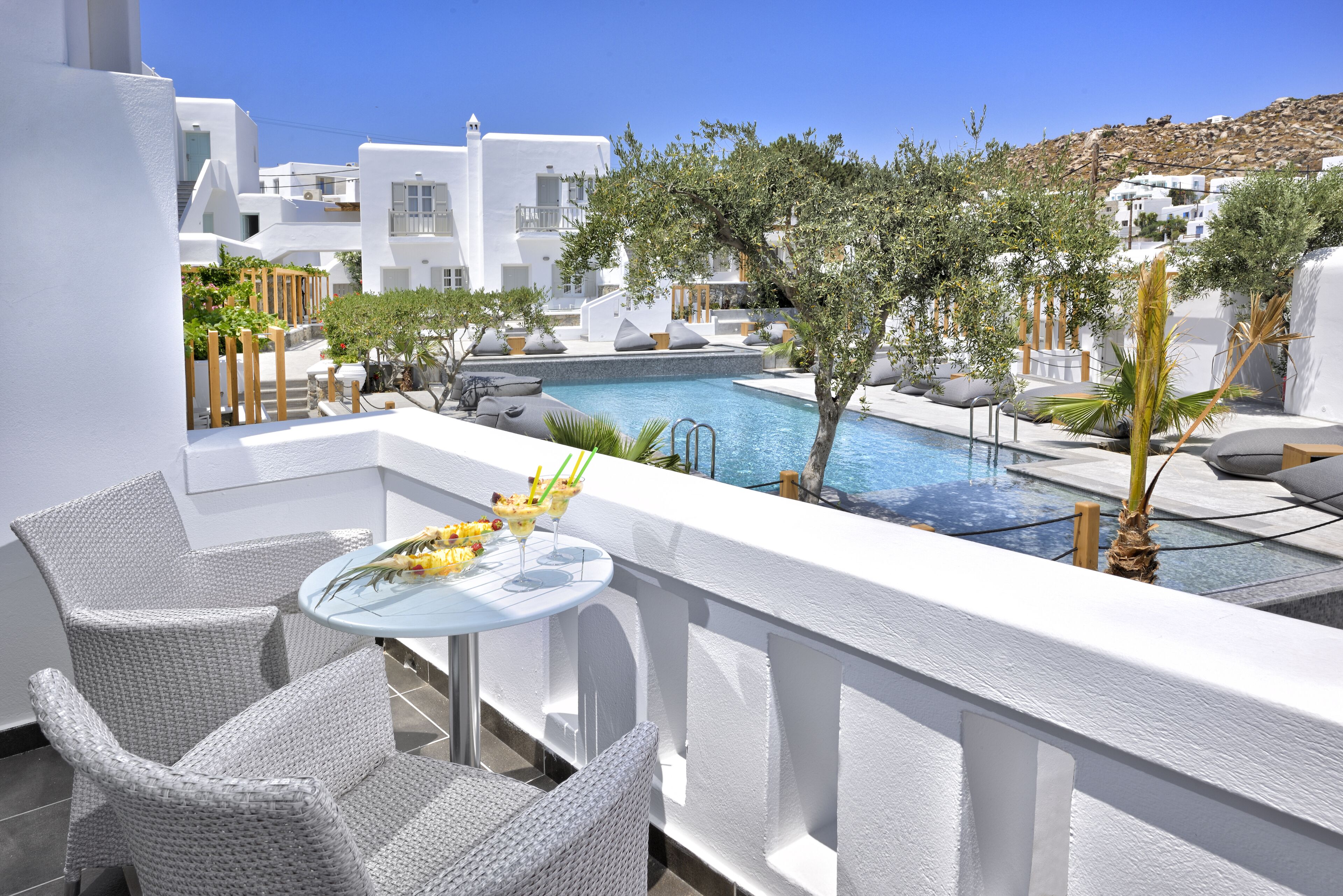 A balcony with wicker chairs, a small round table with a drink, and a pool area with lounge chairs and a tree.
