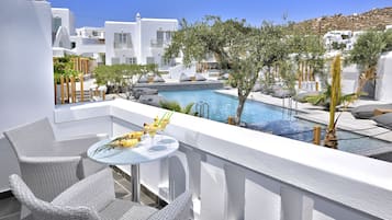 A balcony with wicker chairs, a small round table with a drink, and a pool area with lounge chairs and a tree.