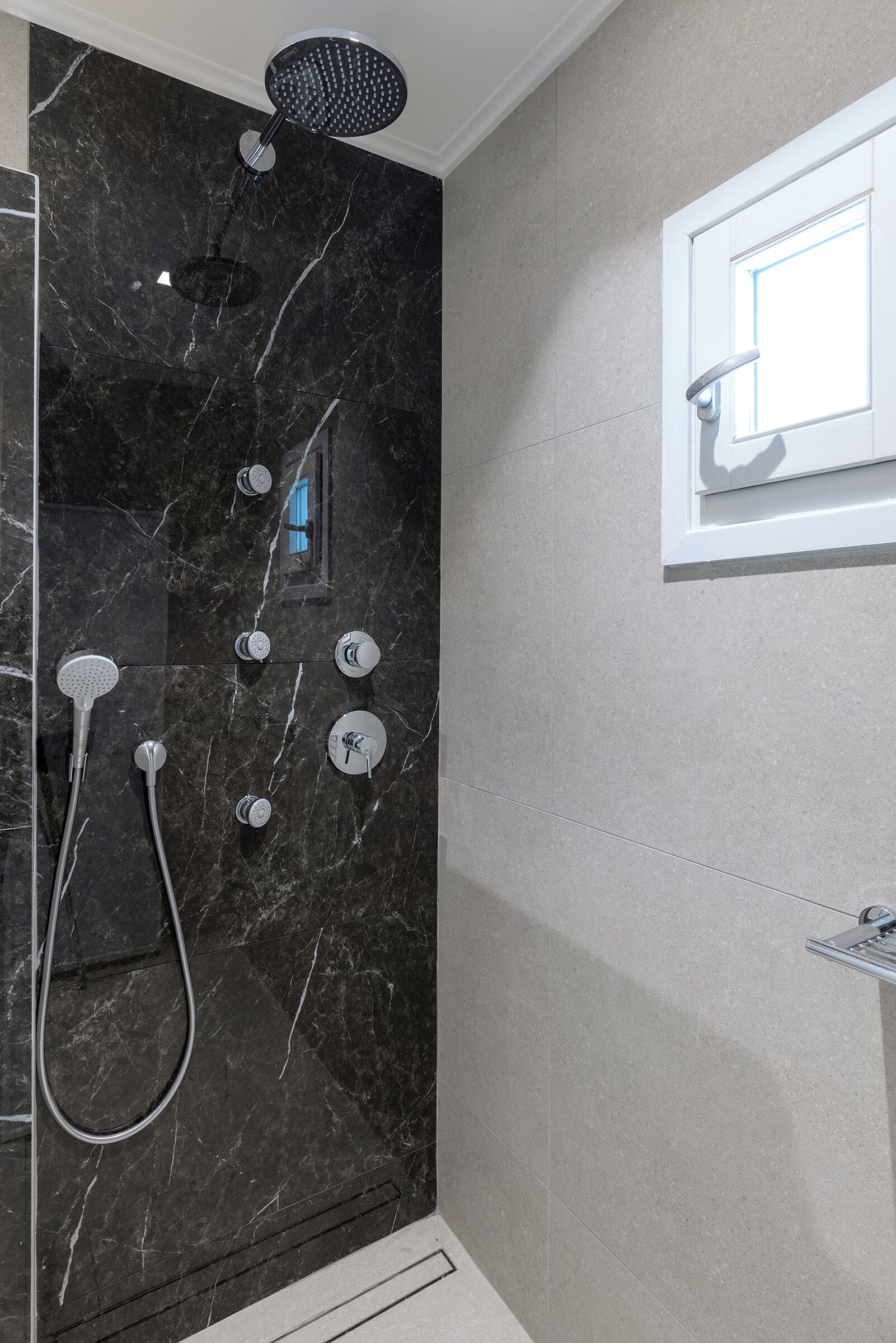 Ein modernes Badezimmer mit schwarzem Marmor an der Duschwand, einer Handbrause, einem wandmontierten Wasserhahn und einem kleinen Fenster über dem Spiegel.