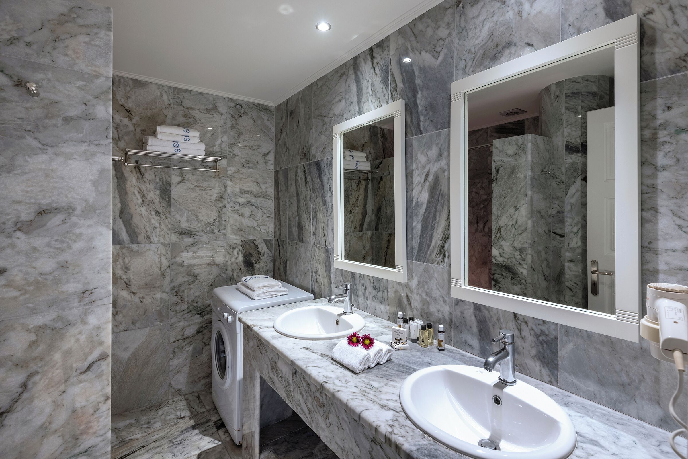 Un baño moderno con paredes de mármol, dos lavamanos, un espejo y lavadora integrada.