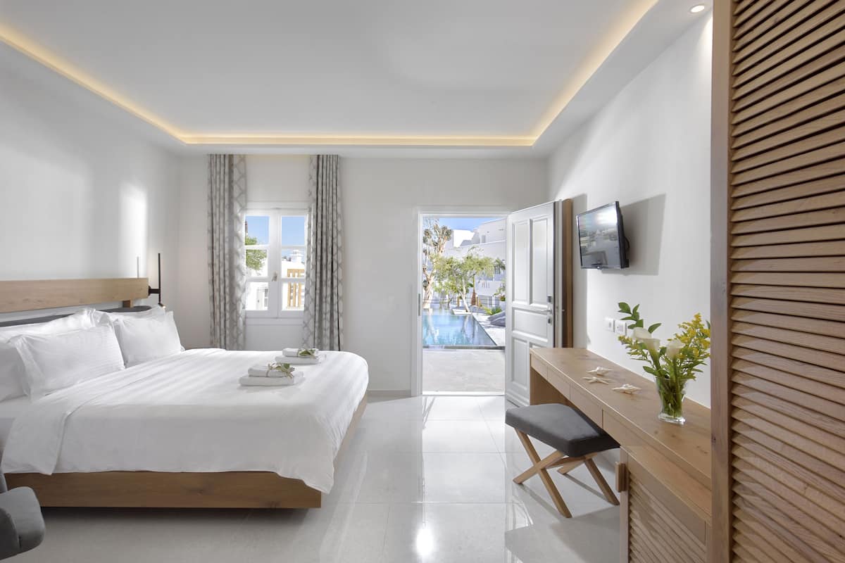 A modern hotel room with a large bed, a desk, a TV, and a view of an outdoor swimming pool.