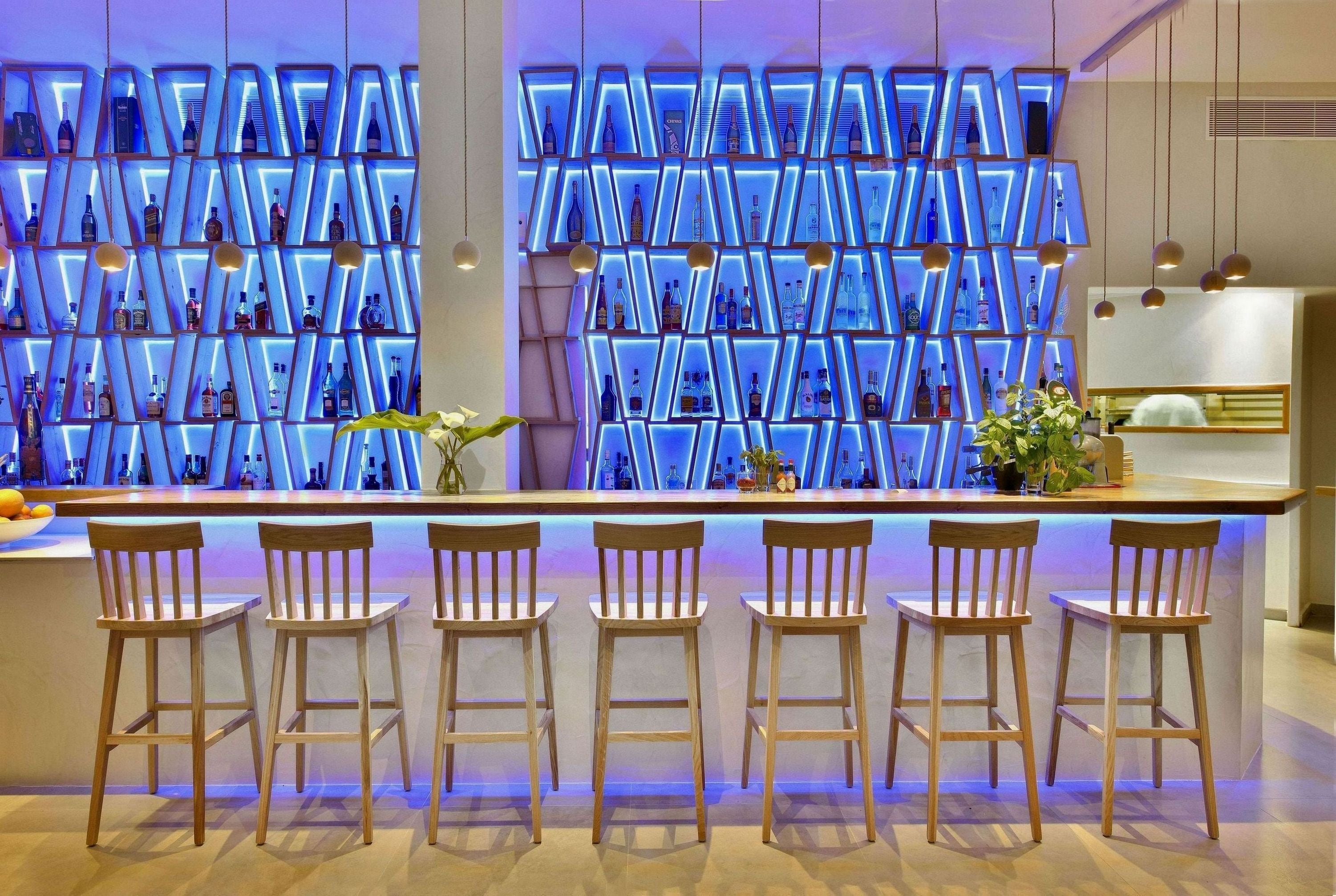 A bar area with a modern design, featuring a wall of illuminated shelves displaying bottles and a bar counter with wooden stools.