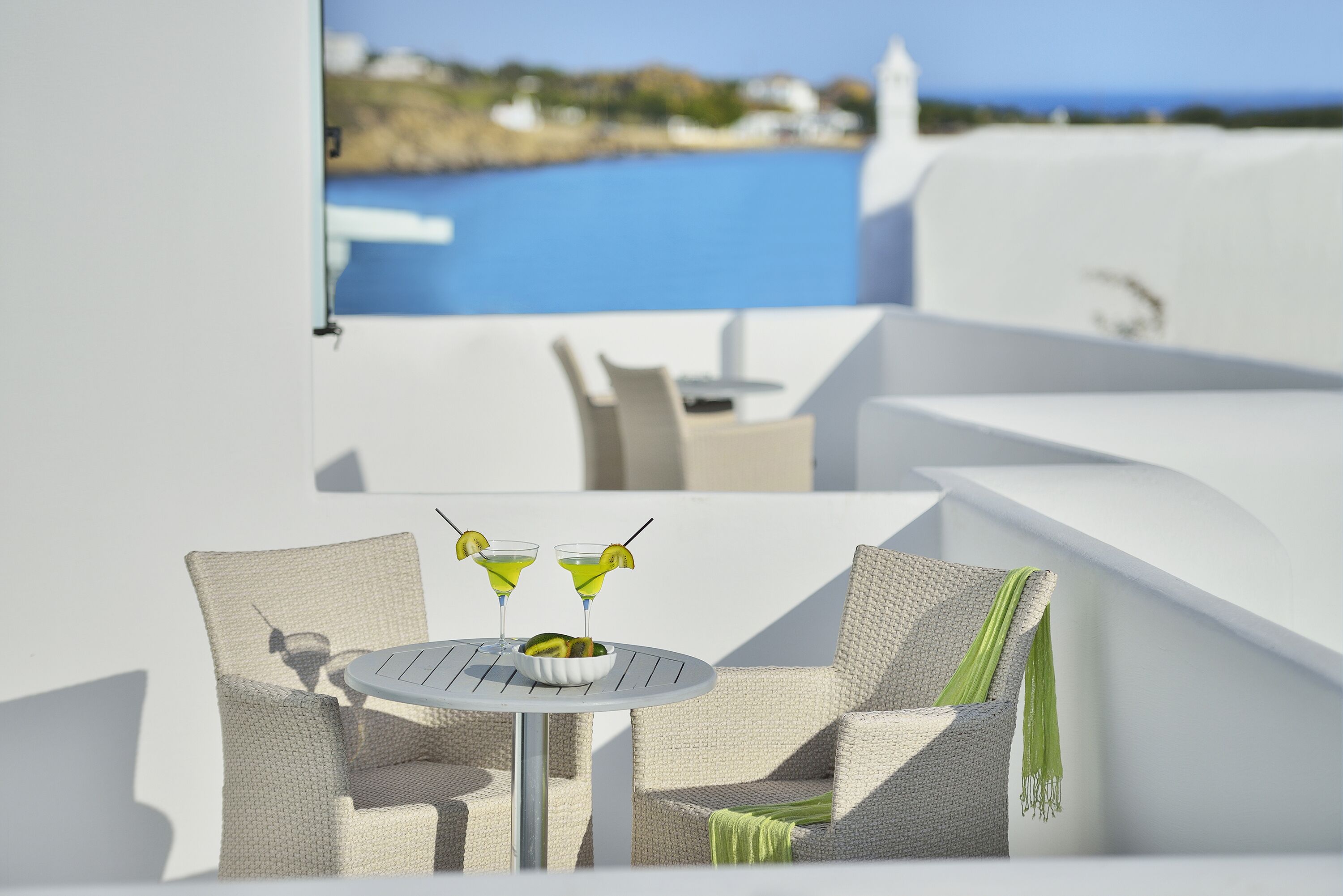 A white outdoor seating area with a round table and two chairs, a small bowl with a fruit, and two martini glasses with green garnishes.