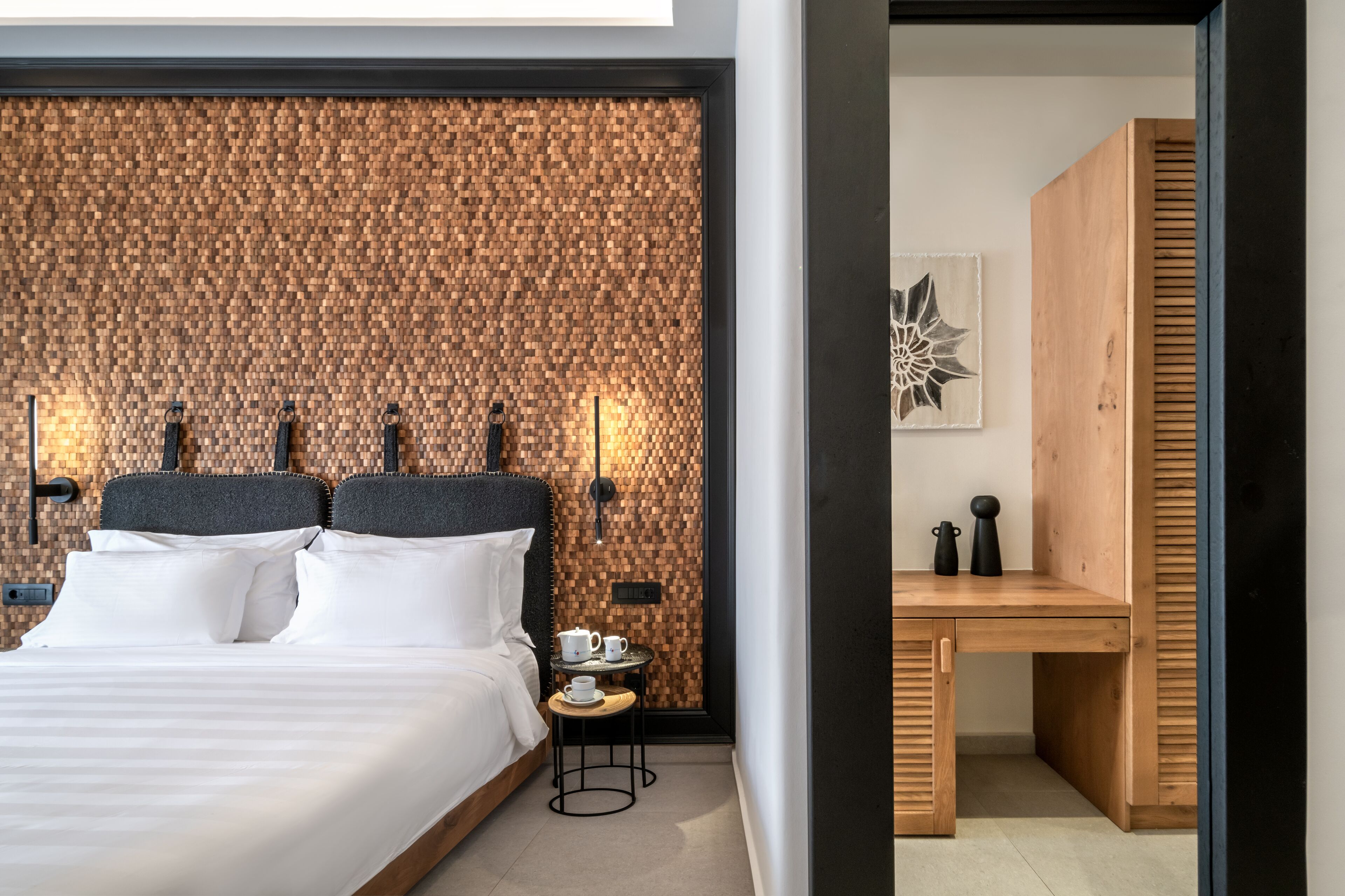 Une chambre d’hôtel moderne avec une tête de lit en bois, une petite table ronde sur laquelle se trouvent une théière blanche et des tasses, et un banc en bois.