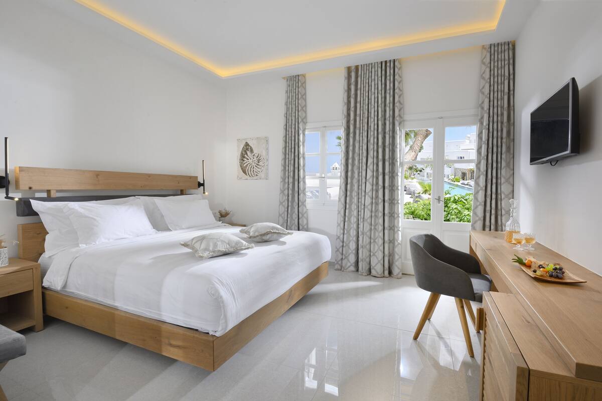 A modern hotel room with a wooden bed, a desk with a fruit plate, a TV, and a view of the pool and greenery.