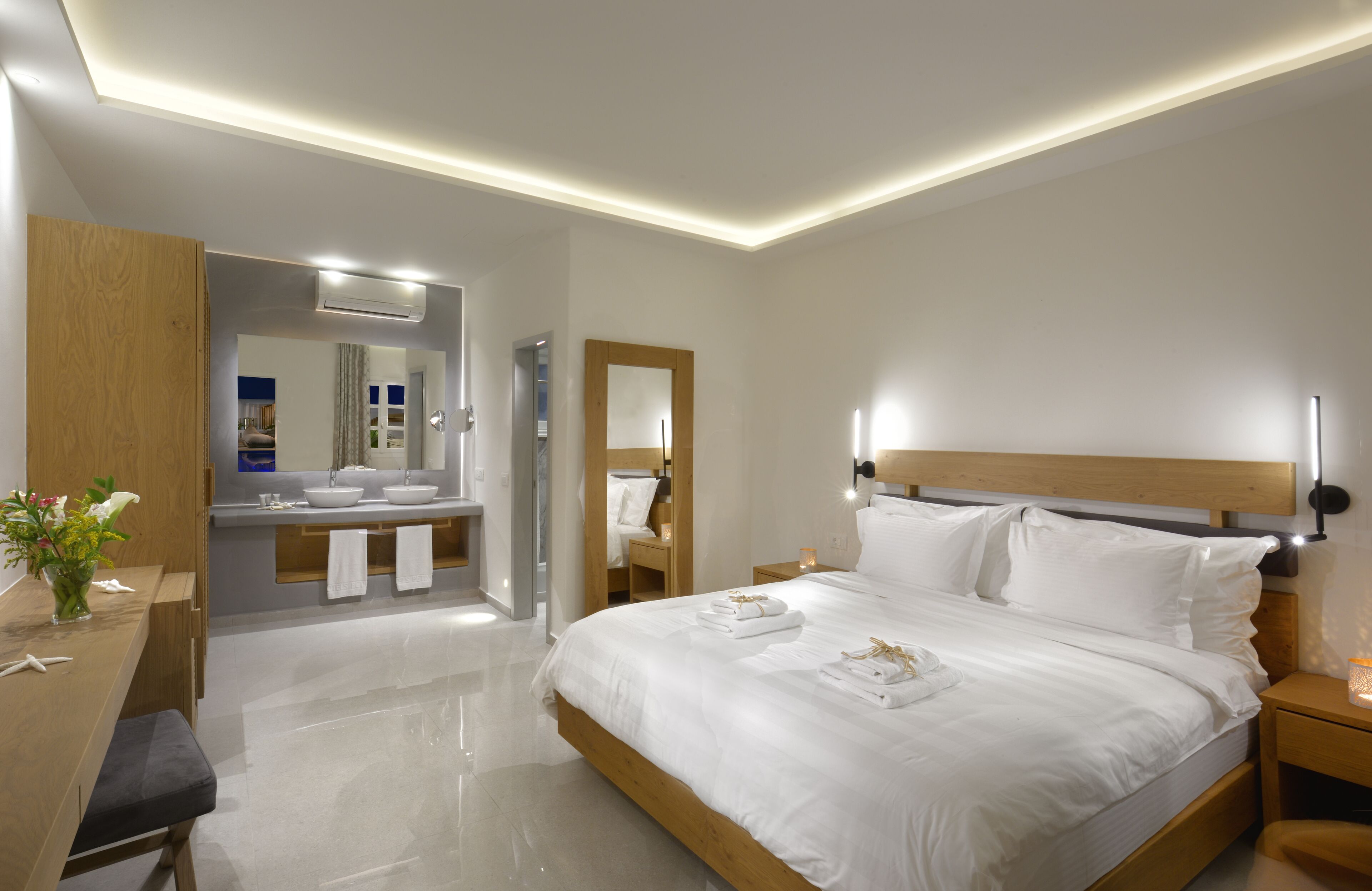 Ein modernes Hotelzimmer mit einem großen Bett, Nachttischen, einem Holzkopfstück, einem Schminktisch mit Spiegel und einem durch eine offene Tür einsehbarer Badezimmer.