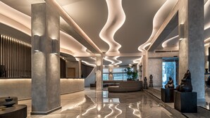 A spacious lobby with a modern design, featuring a curved reception desk, seating areas, and a large window providing natural light. - Hotel Petinos (Mykonos)