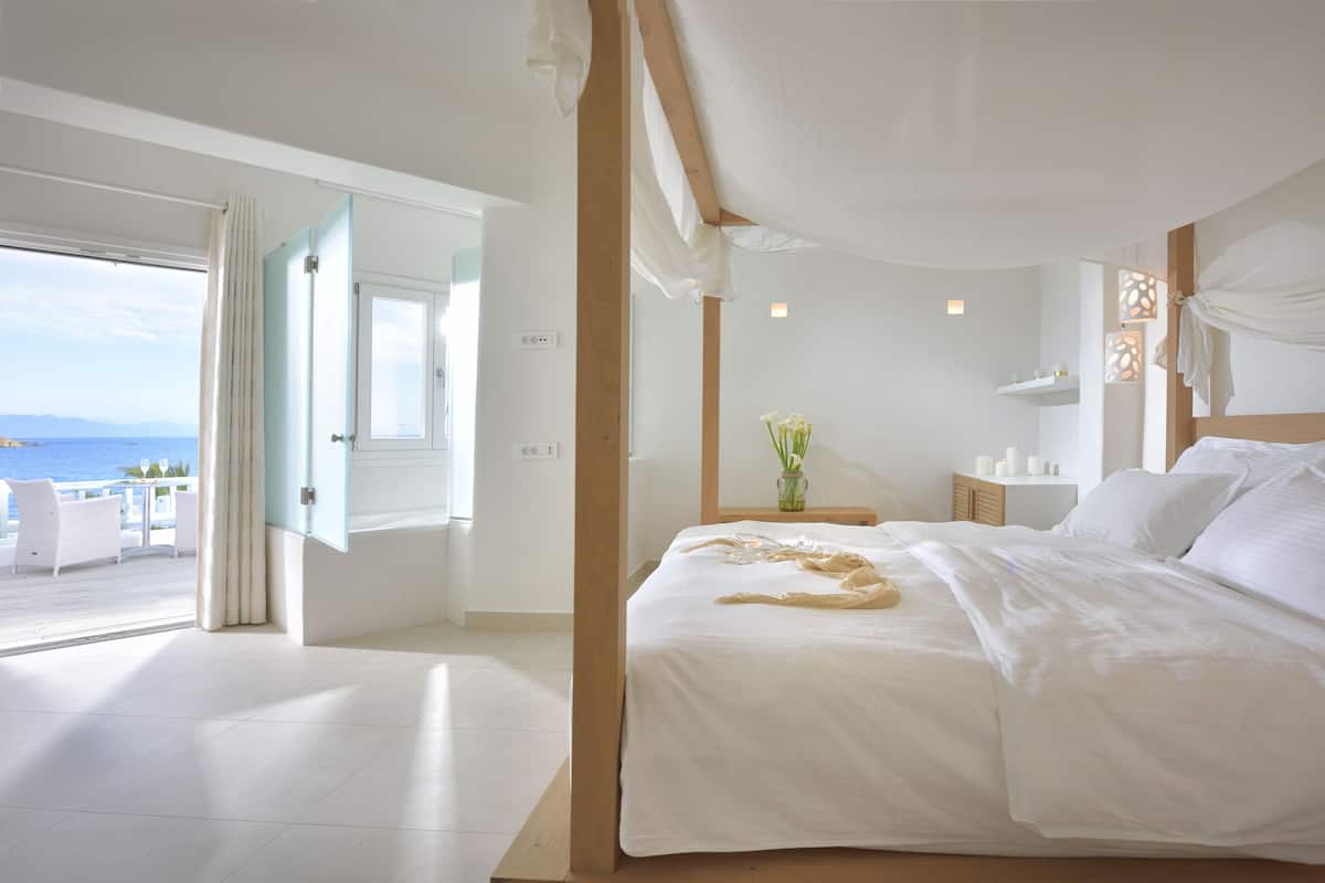 A bedroom with a large bed, a bedside table with a vase of flowers, a view of the sea through an open door, and a balcony with white furniture.
