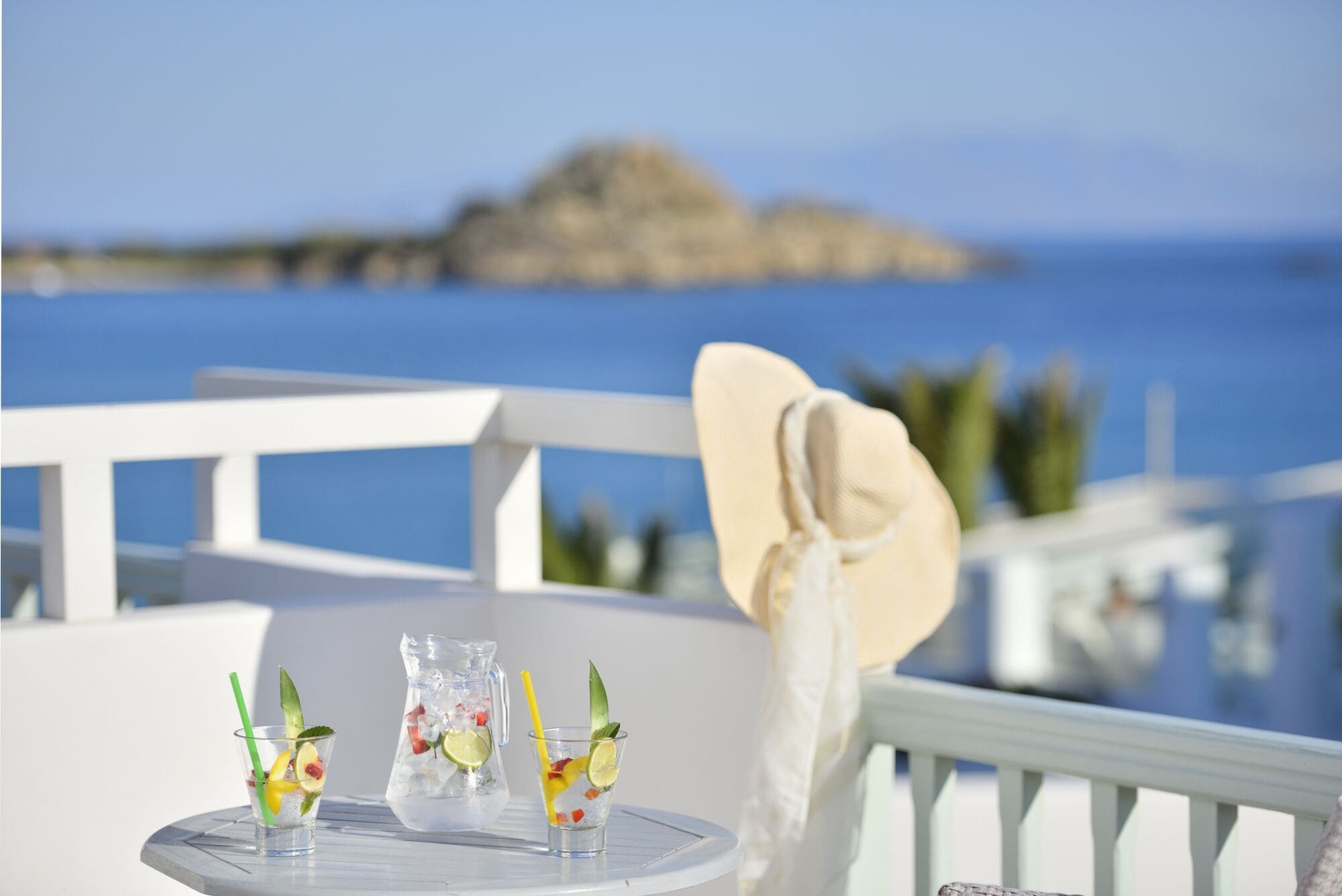 A table with three glasses of iced drinks, a pitcher, and a white hat on a balcony overlooking the sea.