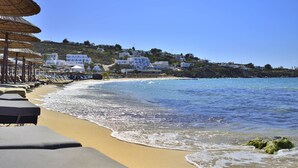 A beach with sun loungers and umbrellas, a clear blue sea, and white buildings on a hillside. - Hotel Petinos (Mykonos)