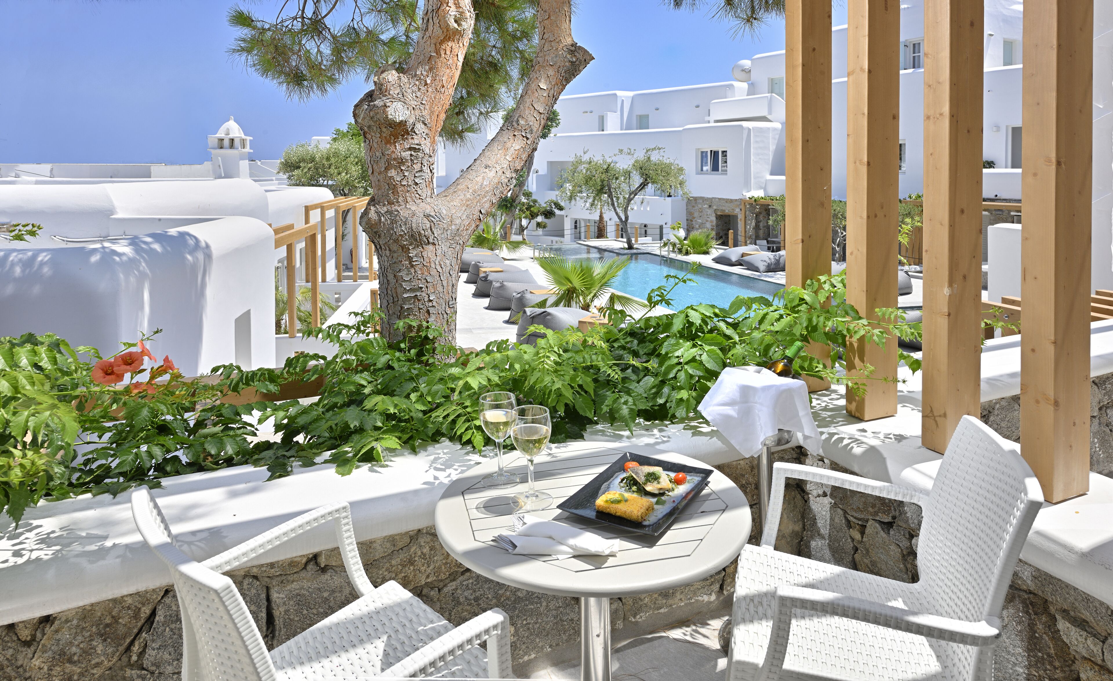 A terrace with white furniture, a table with a meal, and a view of a pool and white buildings.