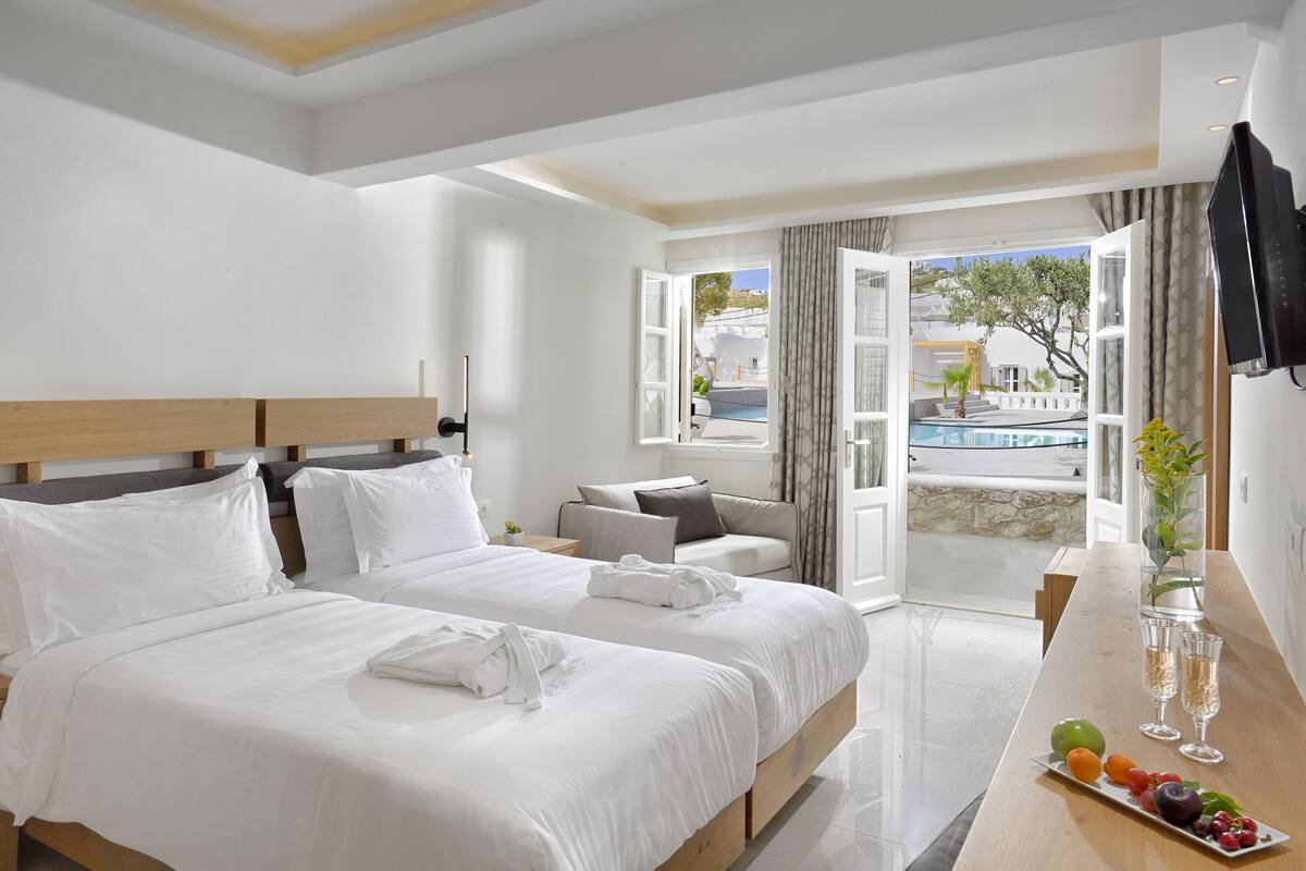 A hotel room with two beds, a small sofa, a dining table with fruit and glasses, and a view of a pool and buildings.