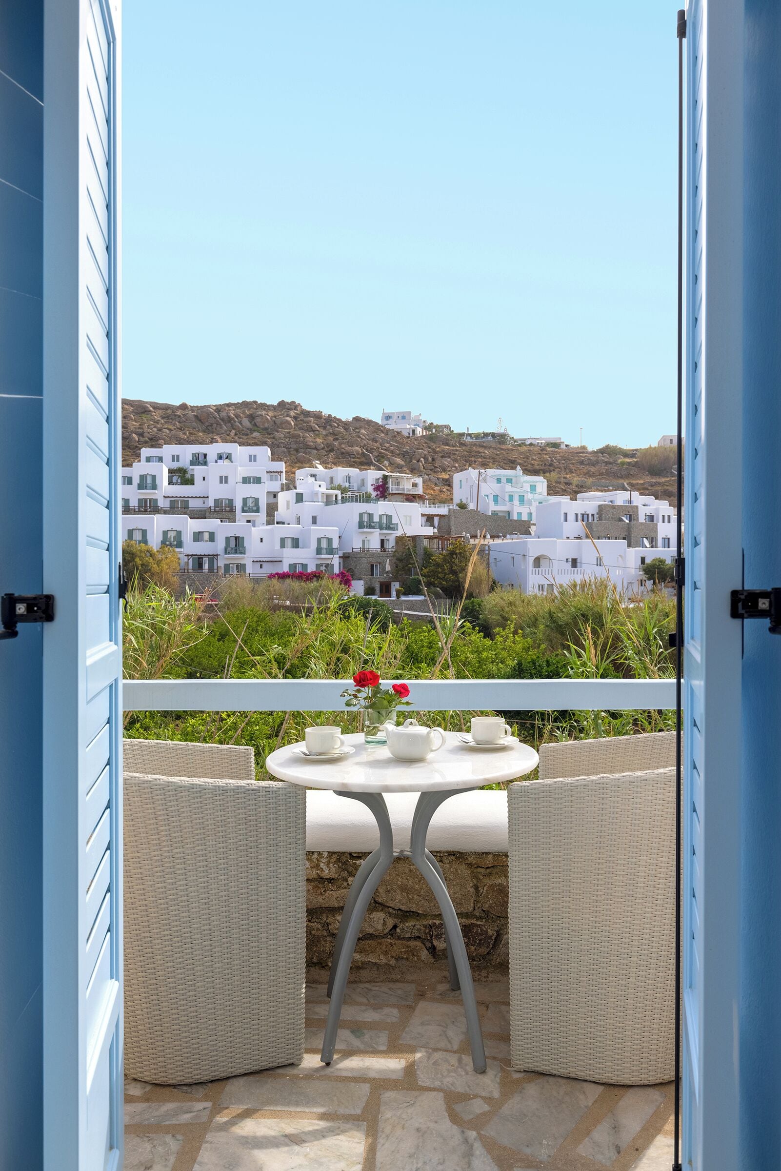 Un balcon avec des fauteuils en osier et une table, d’où l’on peut admirer des bâtiments blancs et une colline.