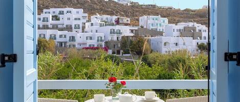 A balcony with wicker chairs and a table, overlooking white buildings and a hillside.
