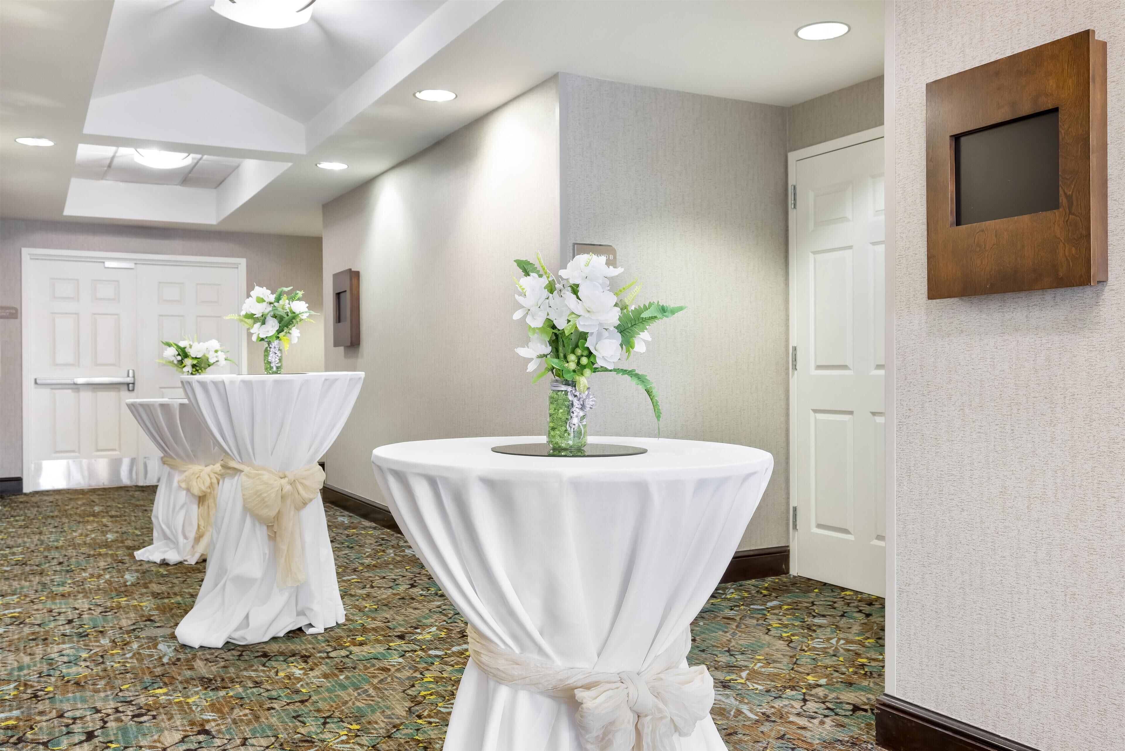 A hallway with round tables covered in white tablecloths and tied with beige sashes, a wall-mounted TV, and a patterned floor.