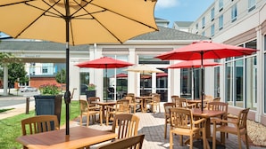 Une terrasse extérieure avec des tables et des chaises en bois, des parasols rouges et une vue dégagée sur la rue en arrière-plan.