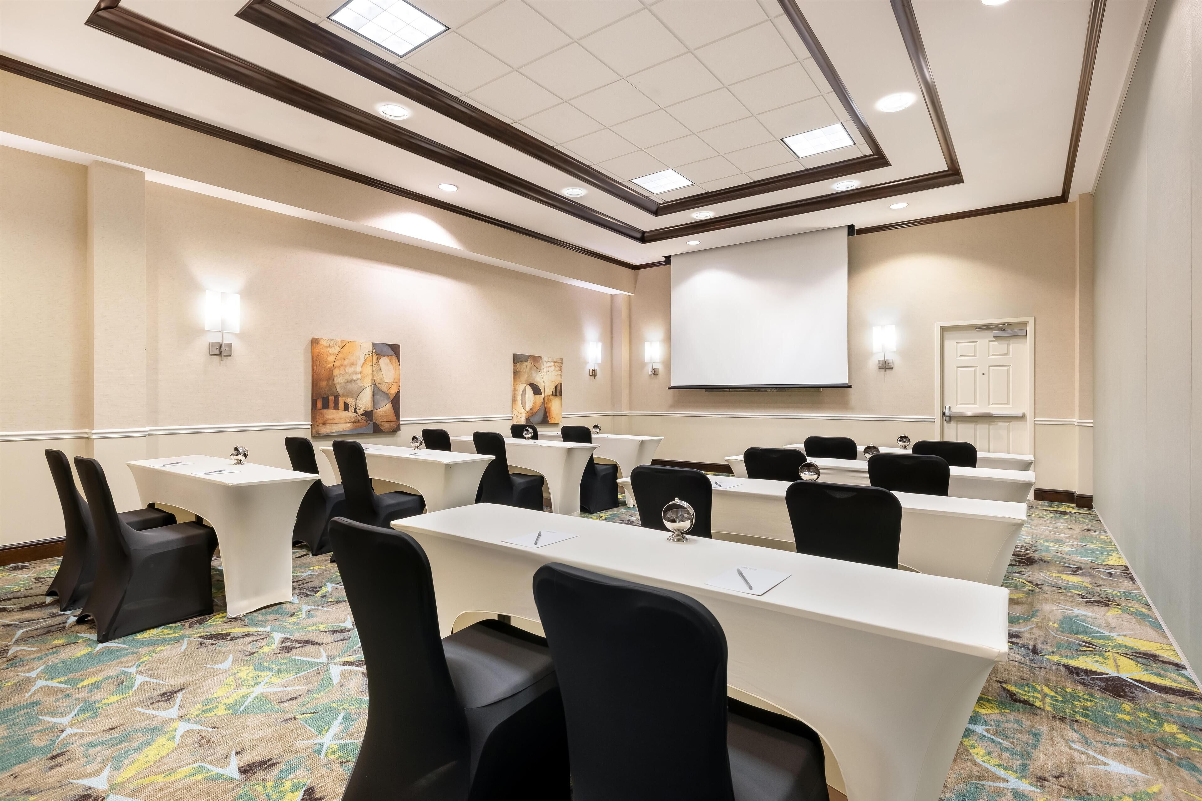 Un salón de conferencias con mesas largas, sillas negras y una pantalla para proyector.