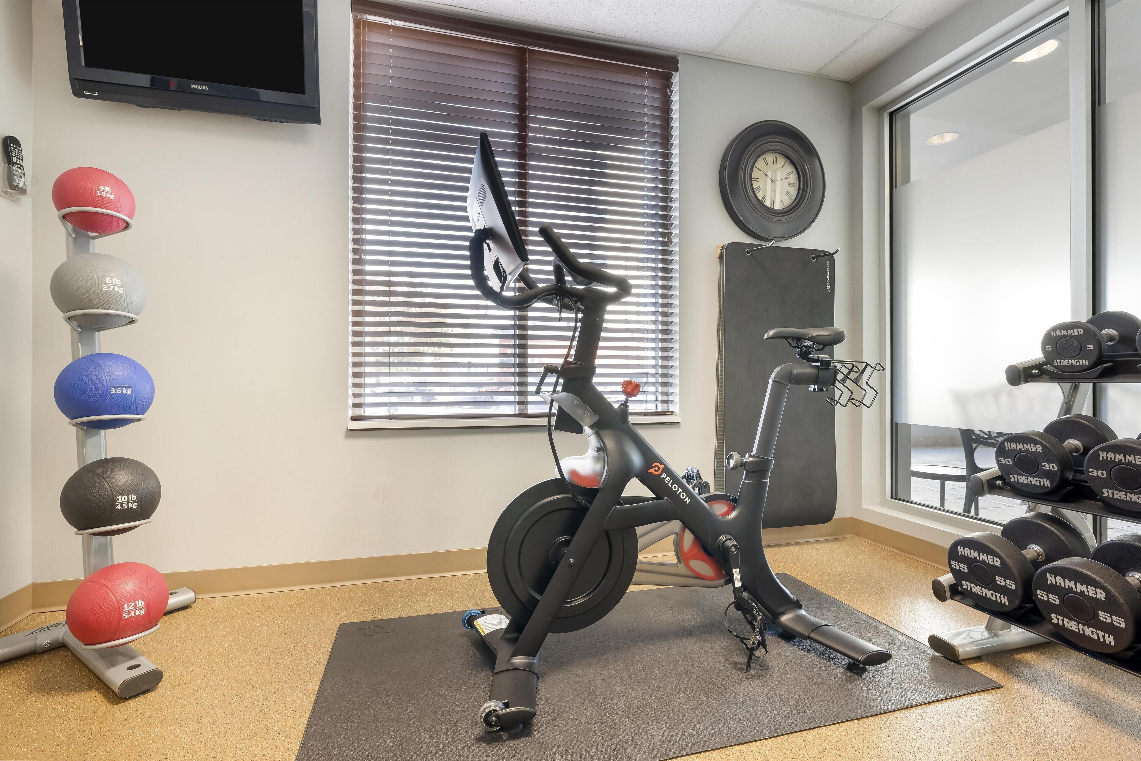 Ein Fitnessraum mit einem Ergometer, einem Wandfernseher, einer Uhr und verschiedenen Gewichten.