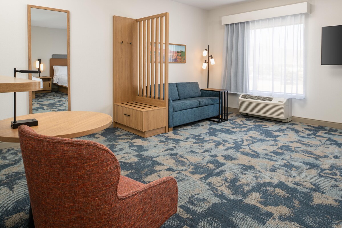 A hotel room with a wooden wardrobe, a blue sofa, a round table, a red chair, a large mirror, a wall-mounted TV, and a window with curtains.